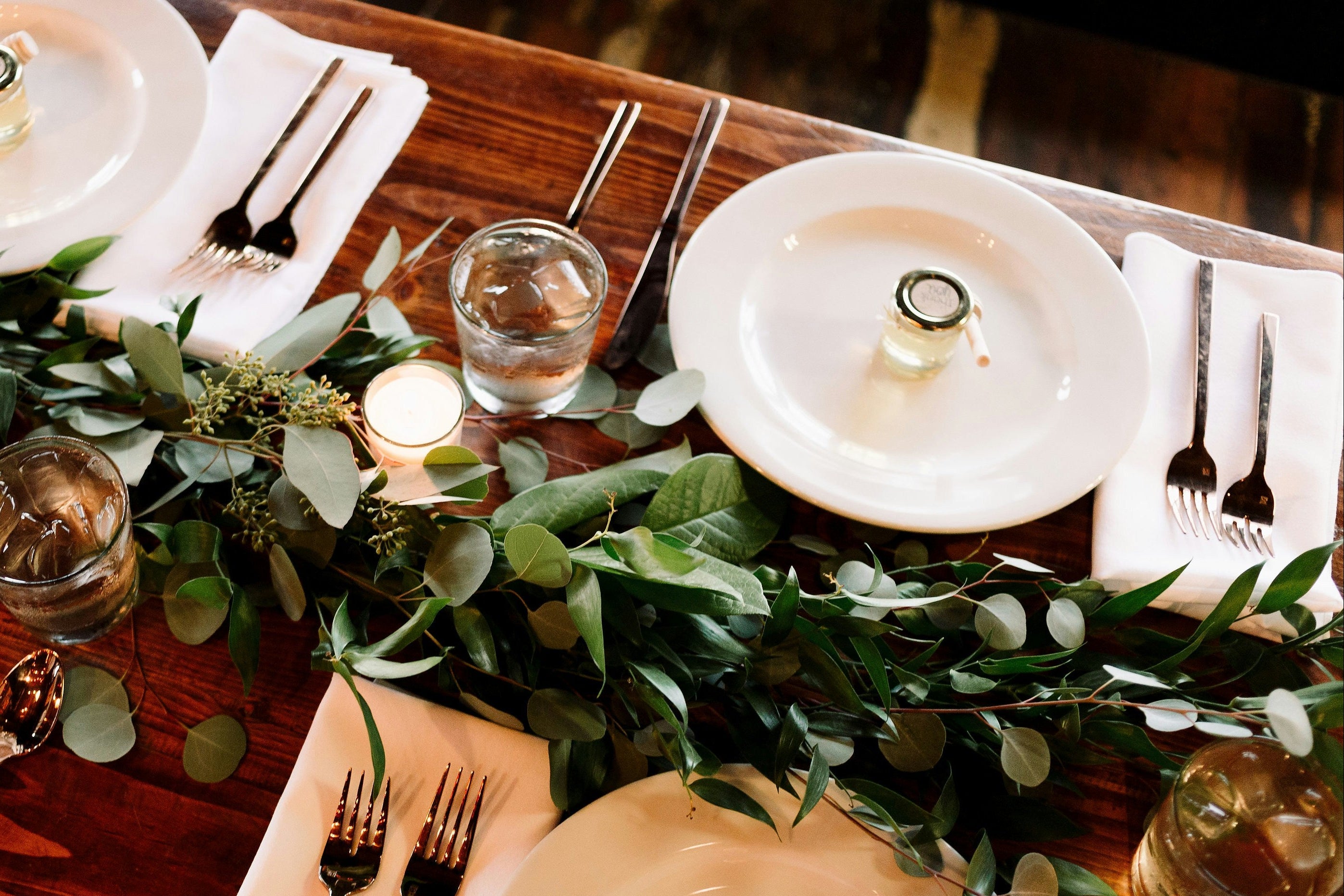 Elegant table setting with white plates, greenery, and candles on a wooden table.